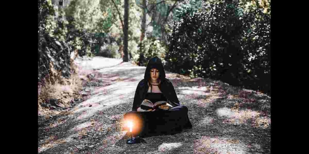 A woman dressed in a black robe sits on the ground with a book, reflecting the Meaning of a Witch in a Dream Biblically.
